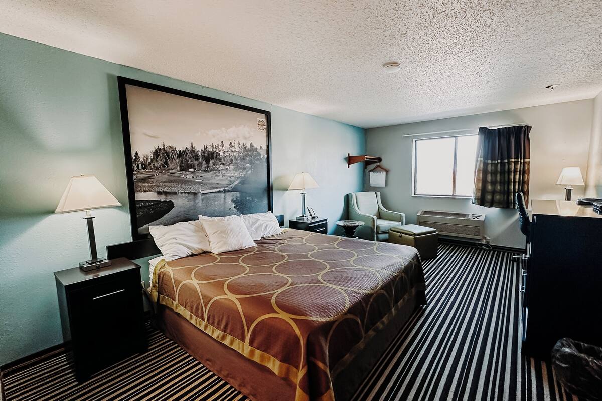 A hotel room with a large bed, two bedside tables with lamps, a chair, a desk with a computer, and a large window with curtains.