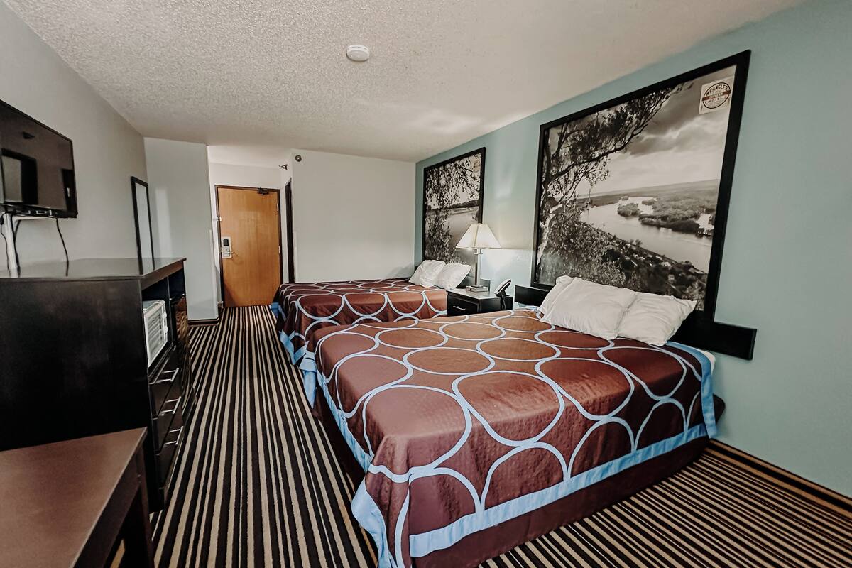 A hotel room with two beds, a desk, a lamp, and a painting on the wall.