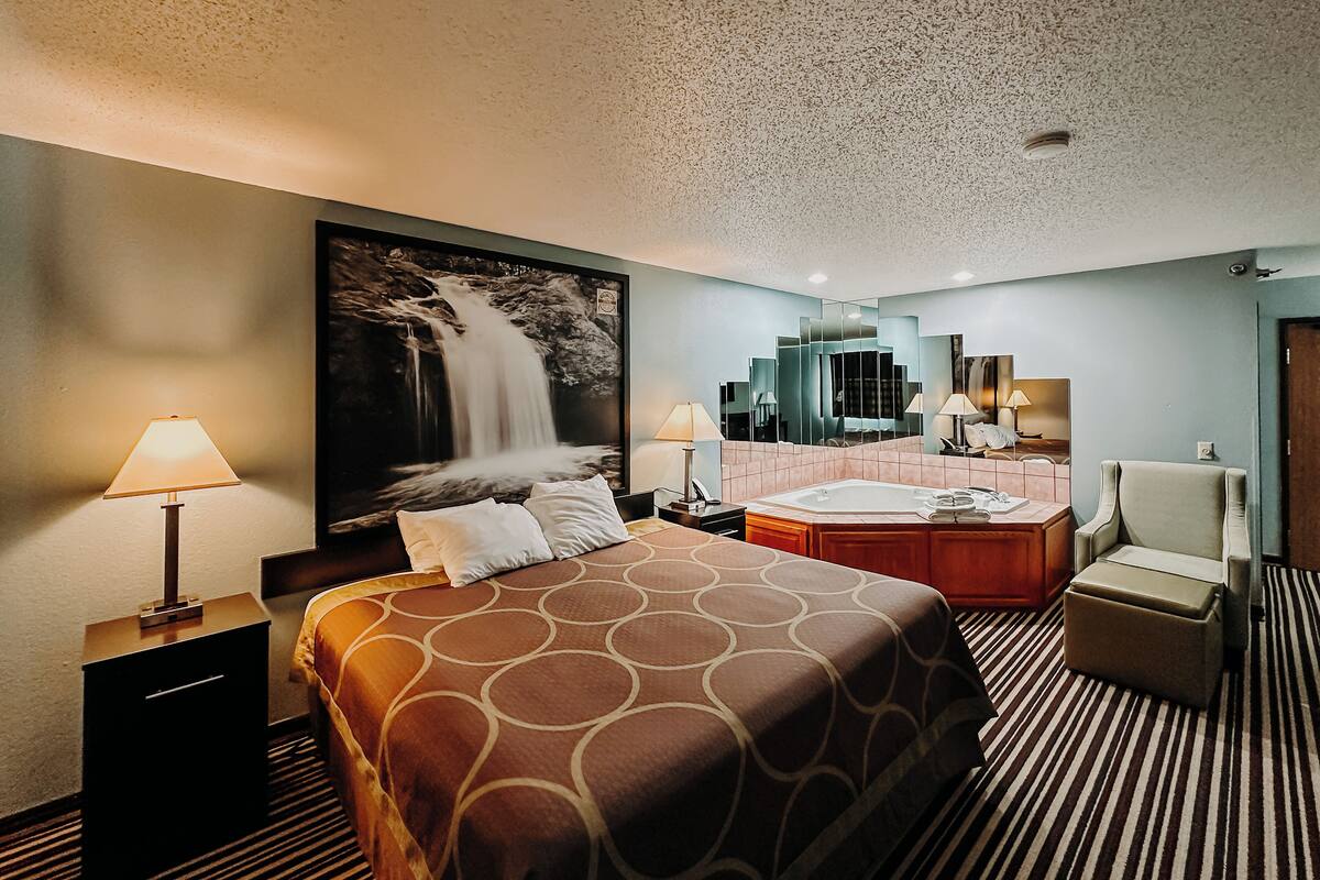 A hotel room with a large bed, a sitting area with a chair, a large mirror, a bathtub, and a waterfall painting on the wall.