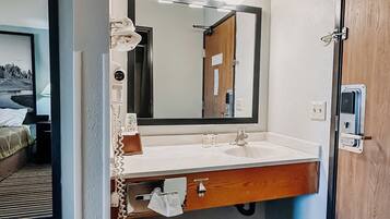 Un bany amb un lavabo, mirall i barra de tovalloles. Hi ha una porta de fusta i un safata de brossa.
