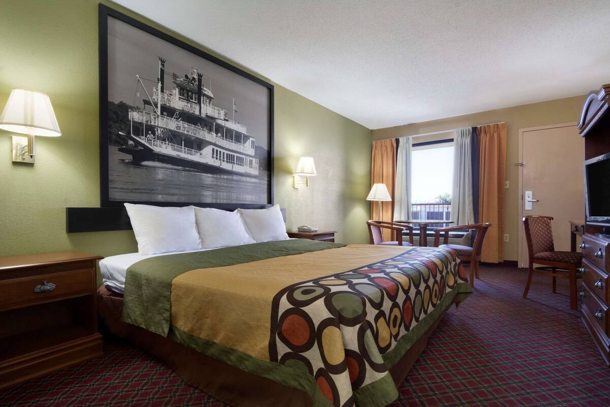 Une chambre d’hôtel avec un grand lit, deux lampes de chevet, un bureau avec une chaise, une télévision et un grand tableau encadré au mur.