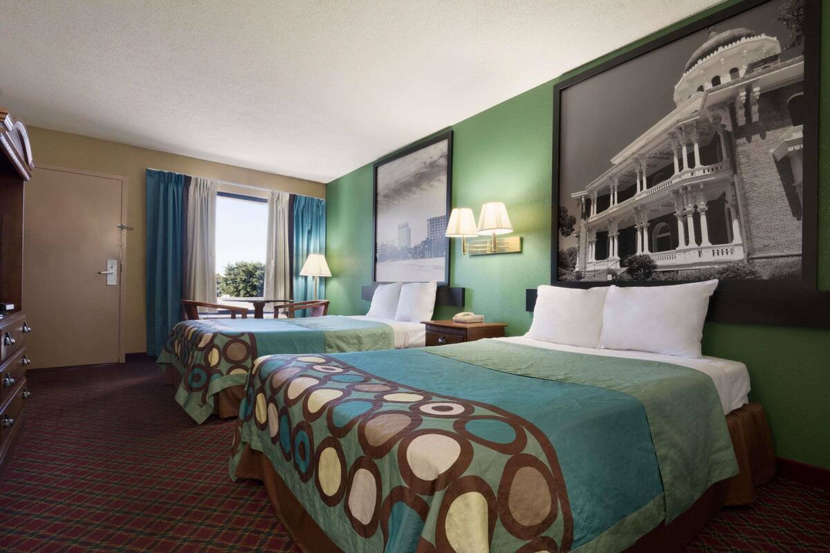 Une chambre d’hôtel avec deux lits, un bureau, une chaise, une fenêtre avec des rideaux et un tableau encadré au mur.