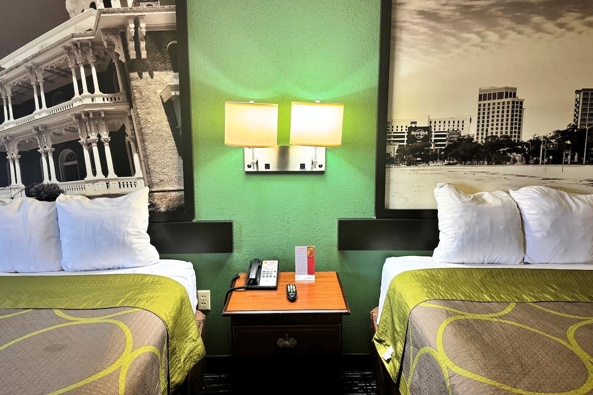 Une chambre d’hôtel avec deux lits, une table de chevet avec un téléphone et une lampe, et un mur vert orné d’une fresque représentant un paysage urbain.