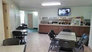 A dining area with tables and chairs, a kitchen area with appliances, and a television on the wall. - Simple Rewards Inn (Guymon)