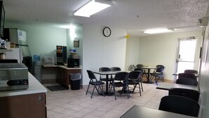 A small, clean room with a kitchenette, a dining area with tables and chairs, a clock on the wall, and a trash bin.