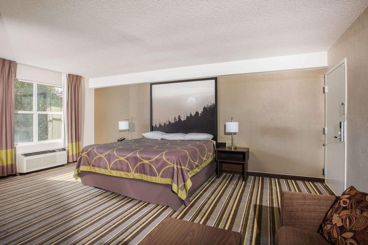 A hotel room with a bed, a nightstand, a lamp, a window with curtains, and a painting on the wall.