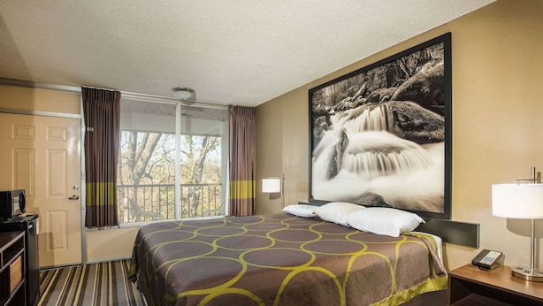 A hotel room with a large bed, a desk, a chair, a window with curtains, and a painting of a waterfall. - Super 8 by Wyndham Sevierville Riverside (Sevierville)