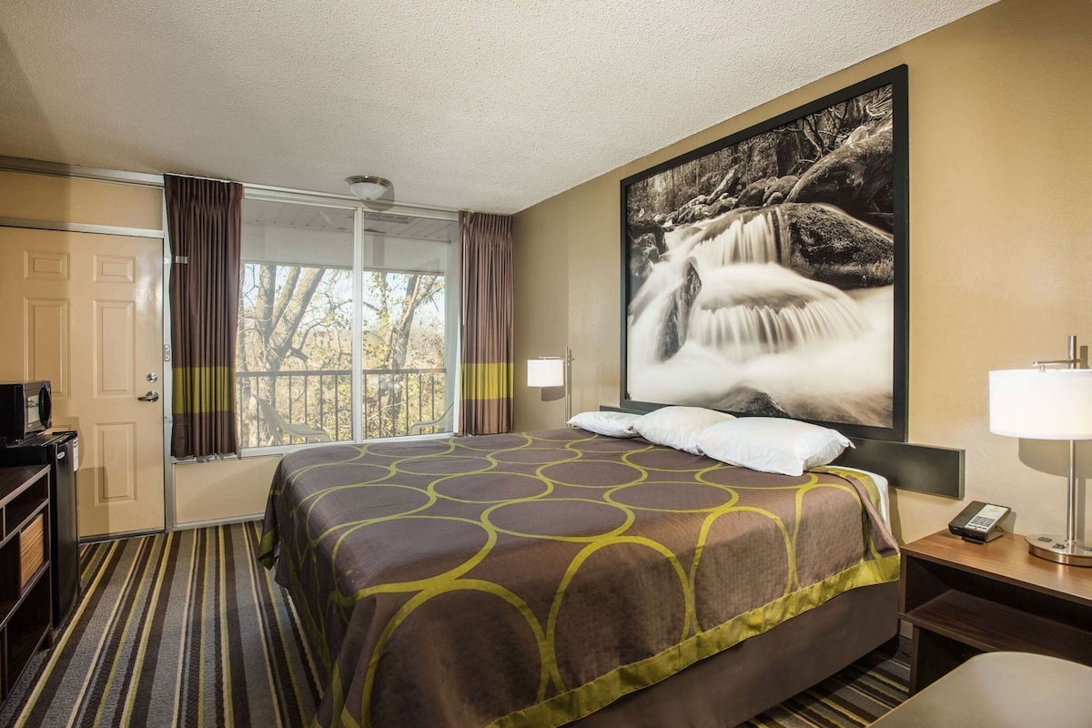 A hotel room with a large bed, a window with curtains, a painting of a waterfall, a bedside table with a lamp, and a telephone.
