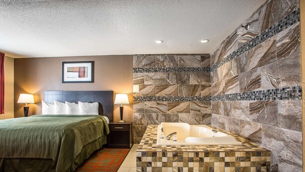 Una habitación de hotel con cama, mesita de noche, lámpara, un espejo grande y baño con bañera y pared de mosaicos.