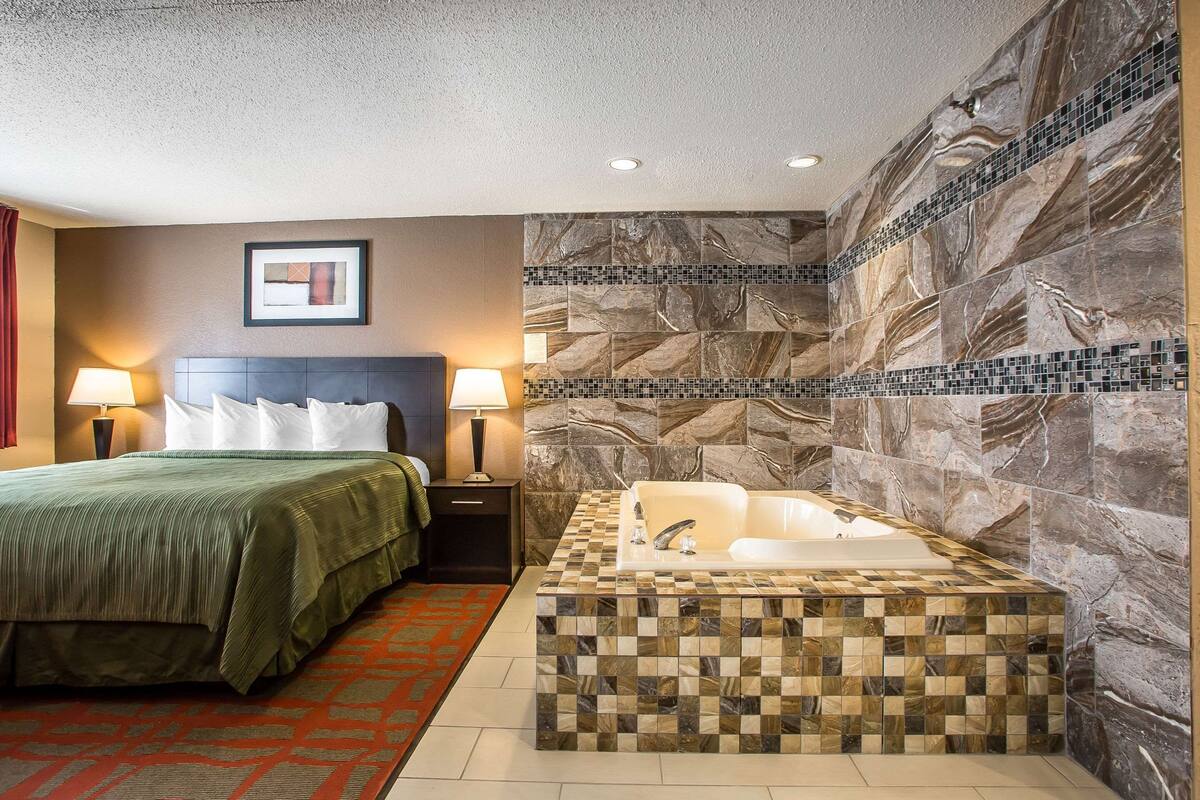 A hotel room with a bed, a nightstand, a lamp, a large mirror, and a bathroom with a bathtub and a mosaic tile wall.