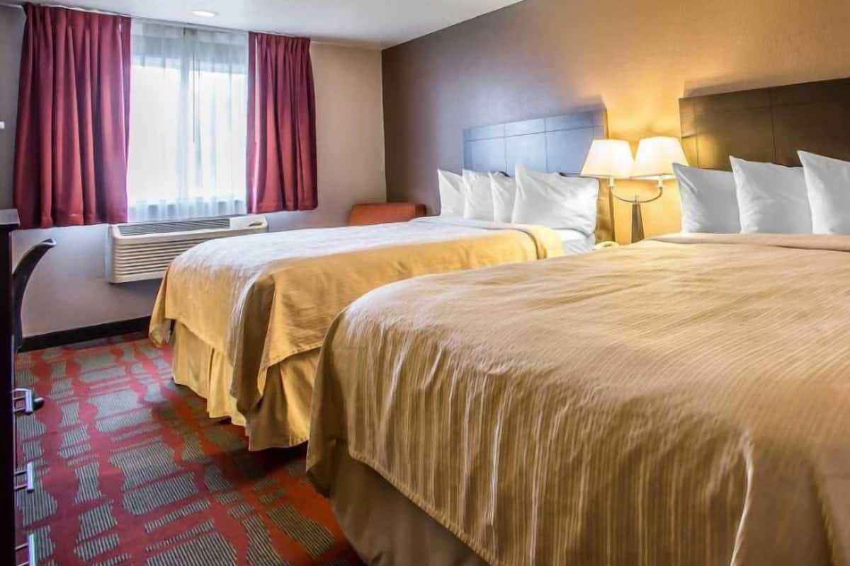 Una habitación de hotel con dos camas, un escritorio, una silla, una lámpara y un aire acondicionado.