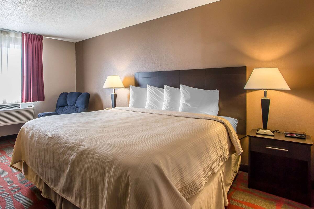 A hotel room with a large bed, two bedside lamps, a chair, and a nightstand with a remote control.
