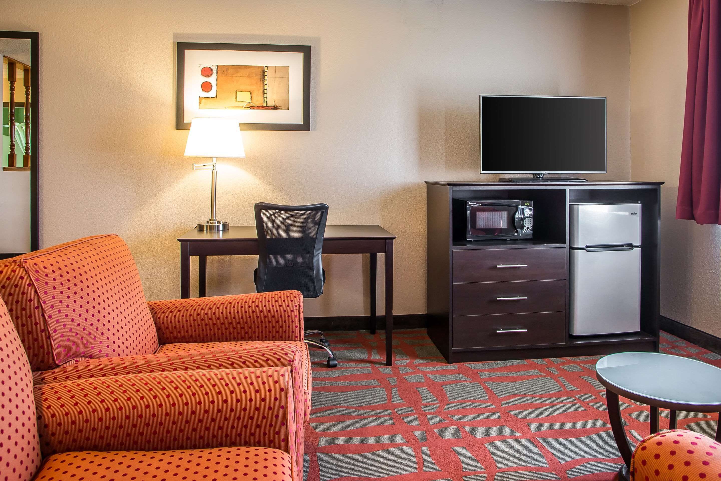 Quarto de hotel com sofá, escrivaninha com luminária, televisão, mini-geladeira e uma pequena mesa.