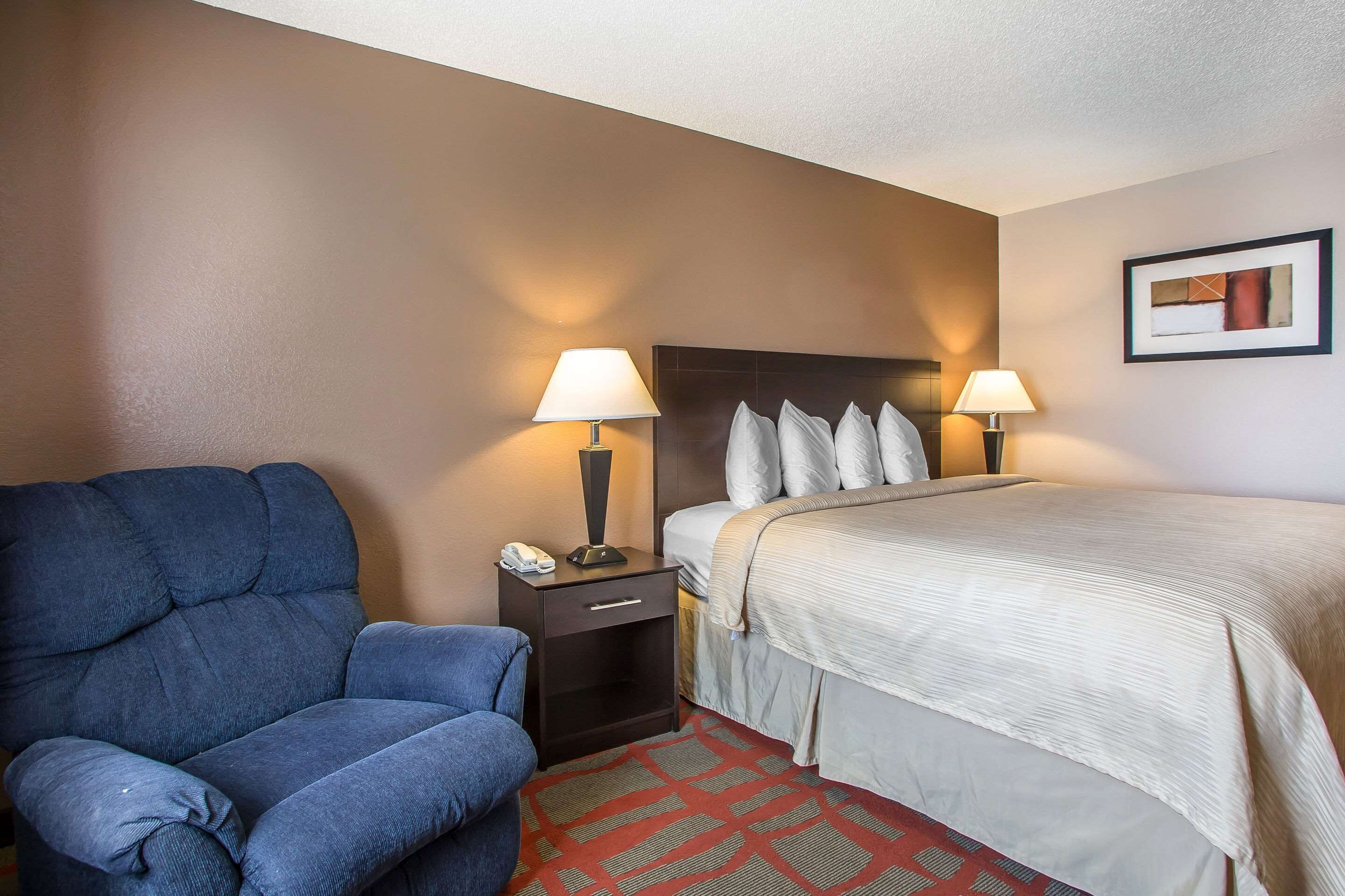 A hotel room with a bed, two bedside lamps, a nightstand with a phone, a blue armchair, and a patterned rug.