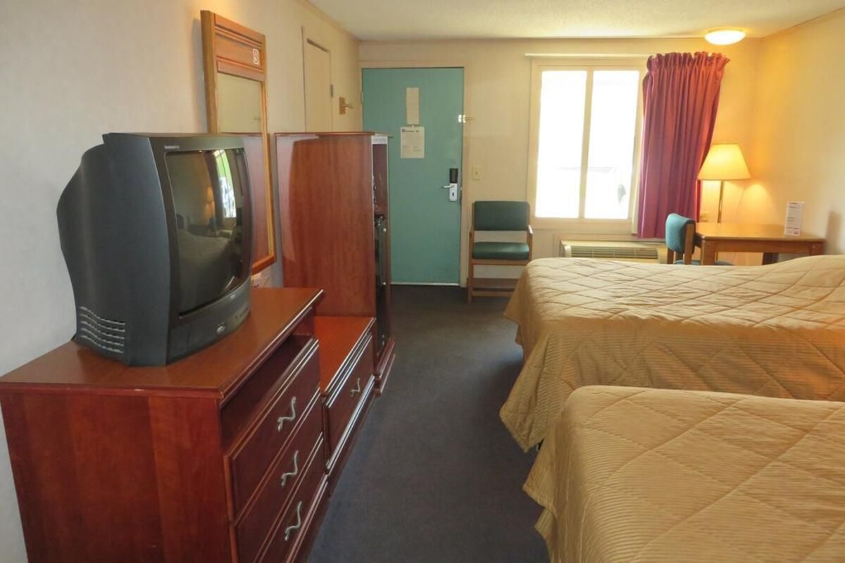Hotelkamer met televisie op een dressoir, een bed met quilt, bureau met lamp en raam met gordijnen.