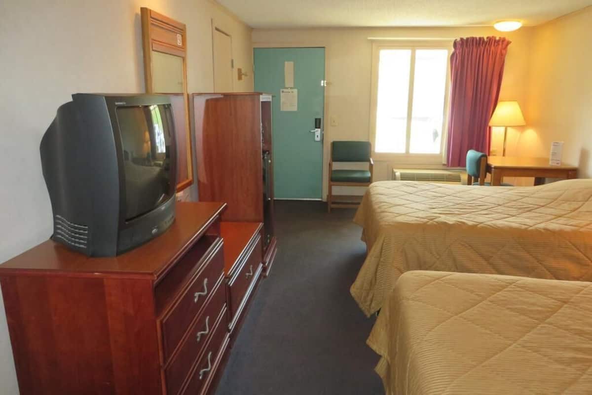 Ett hotellrum med en tv-apparat på en byrå, en säng med ett täcke, ett skrivbord med en lampa och ett fönster med gardiner.
