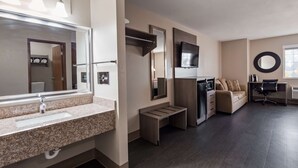Une chambre d’hôtel moderne dotée d’un lavabo avec un plan de travail en granit, d’un grand miroir, d’un téléviseur à écran plat, d’un micro-ondes encastré, d’un petit banc, d’un canapé et d’un miroir rond fixé au mur.
