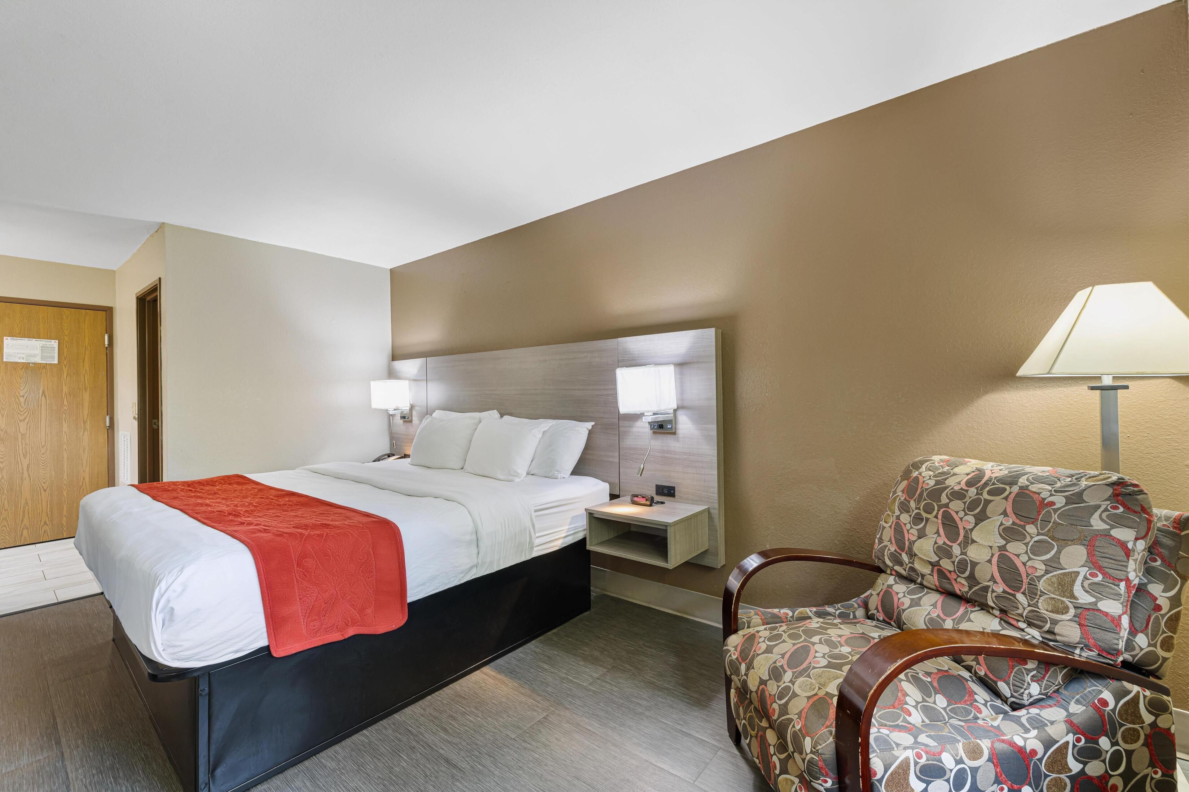 A hotel room with a bed, a nightstand, a chair, and a lamp.