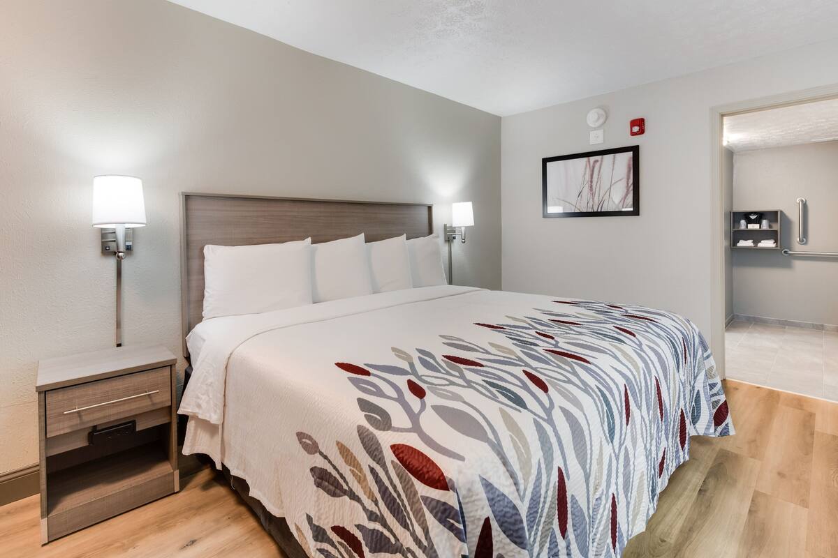 A hotel room with a bed, bedside tables, a nightstand, a wall-mounted lamp, a framed picture, and a bathroom visible through an open door.