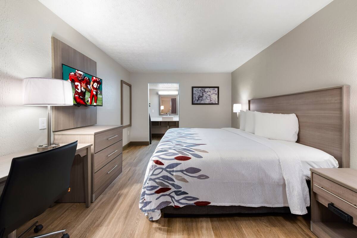 A hotel room with a bed, a desk with a chair, a TV, and a bathroom visible through an open door.
