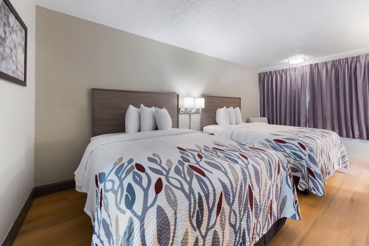 Habitación de hotel con dos camas, cabecera con espejo, lámpara de pared y ventana con cortinas.