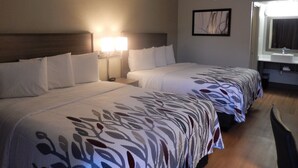 A hotel room with two beds, a nightstand with a lamp, a bathroom with a sink, and a framed picture on the wall.