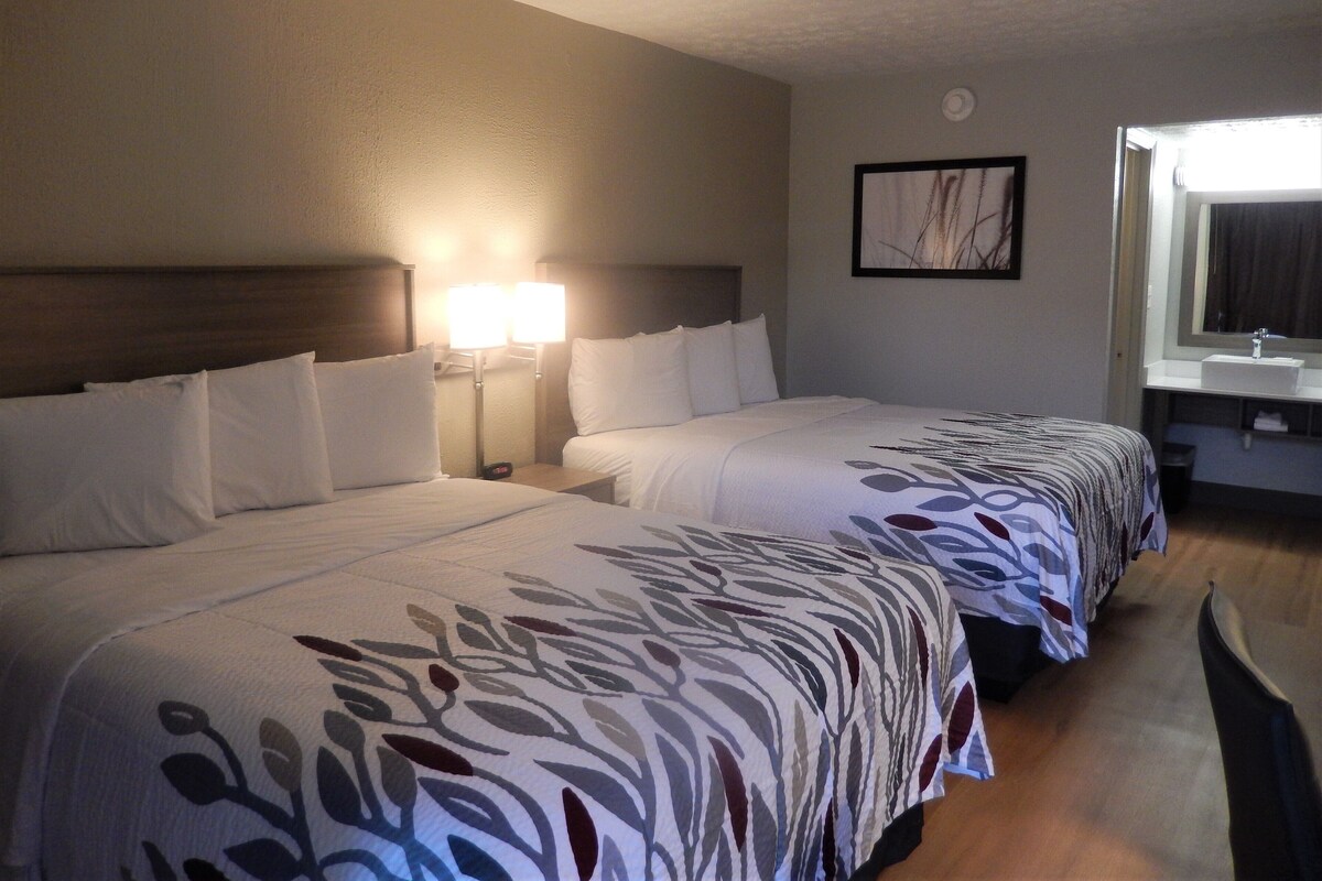 A hotel room with two beds, a nightstand with a lamp, a bathroom with a sink, and a framed picture on the wall.
