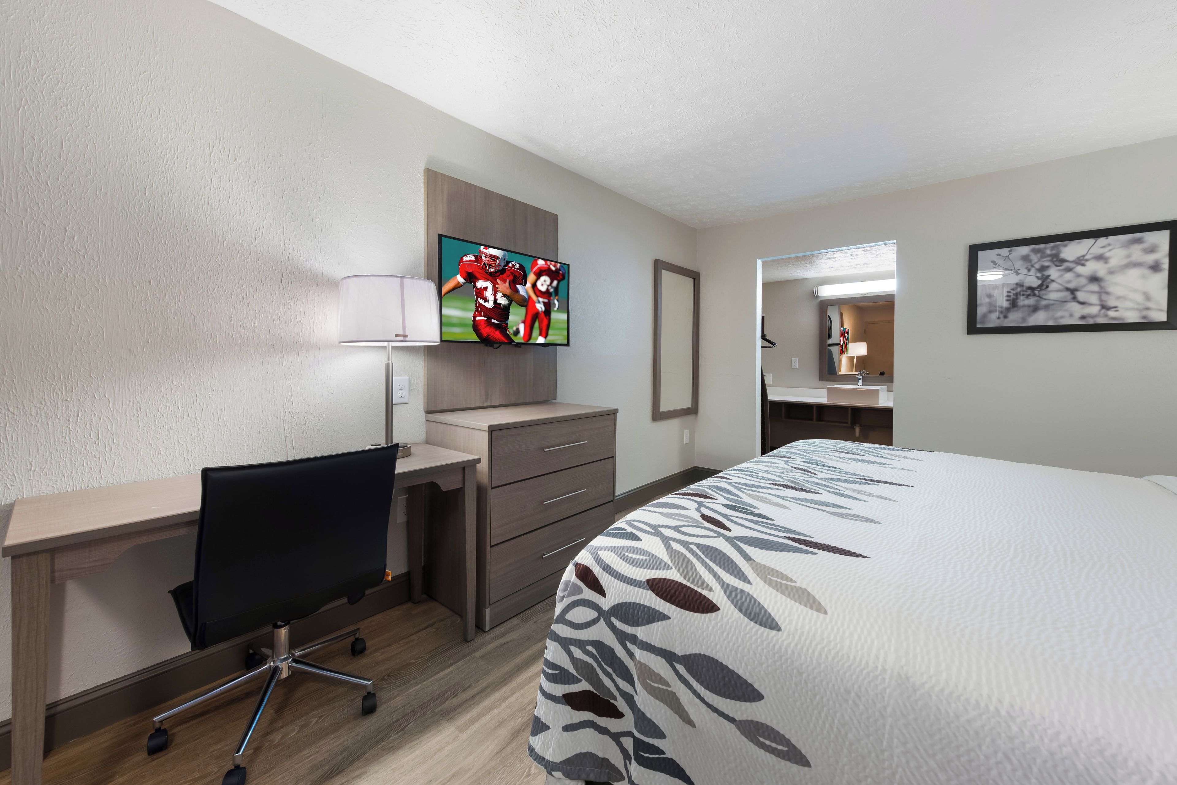 Habitación de hotel con una cama, un escritorio con silla, televisor, espejo y baño visible a través de una puerta abierta.
