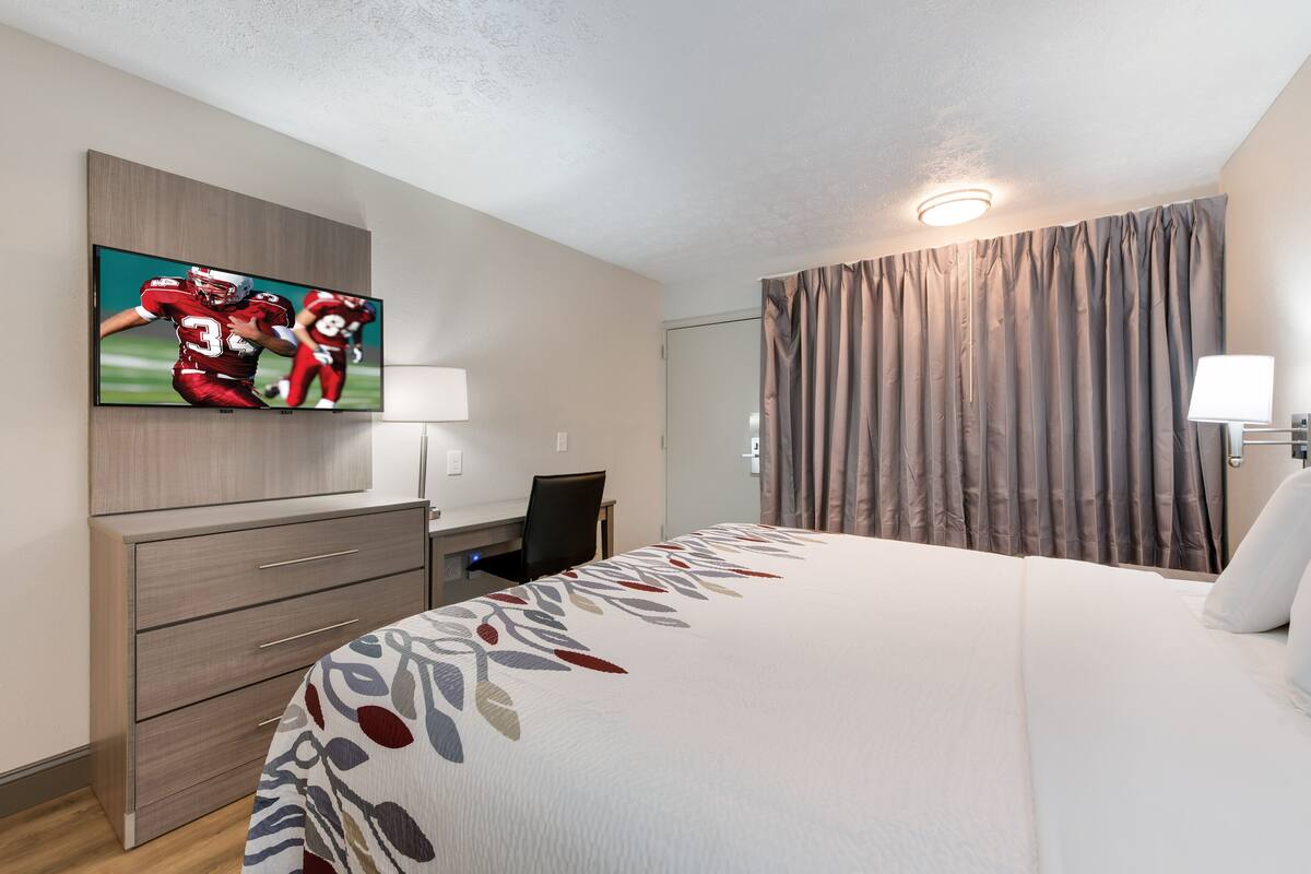 A hotel room with a large bed, a desk with a chair, a flat-screen TV mounted on the wall, and a window with curtains.