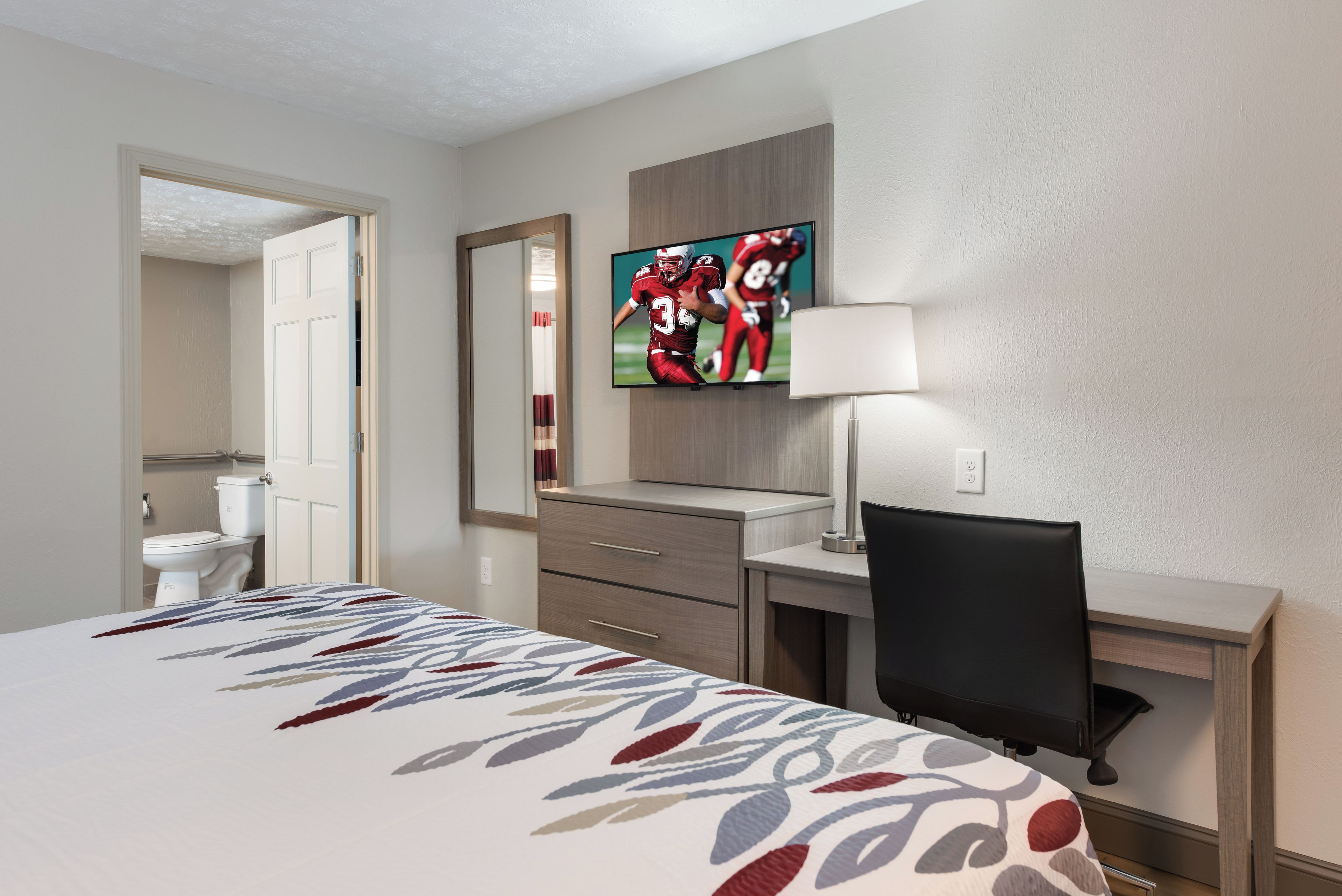 A hotel room with a bed, a desk, a chair, a TV, a mirror, and a bathroom visible through an open door.