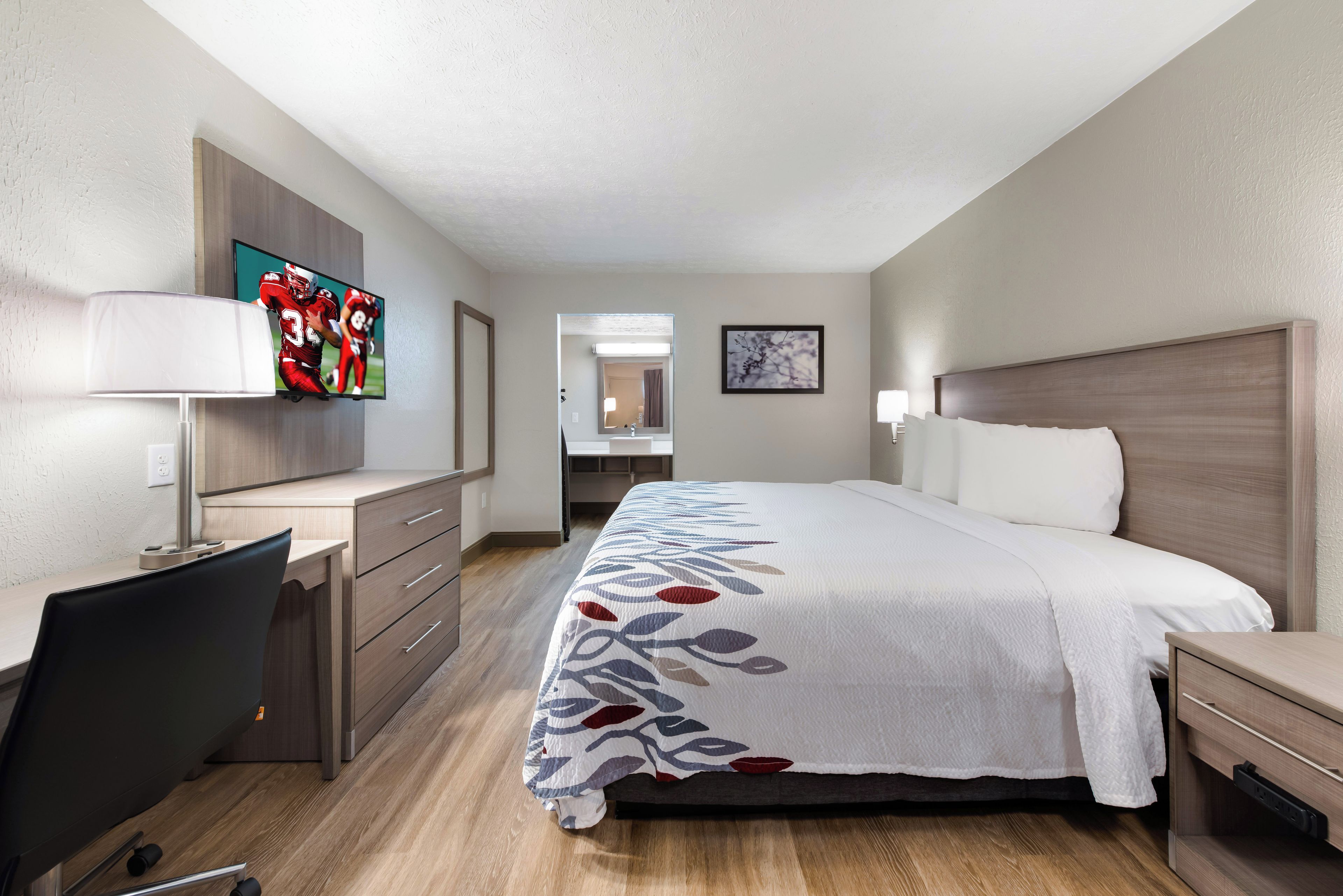 A hotel room with a bed, desk, chair, TV, and a bathroom visible in the background.