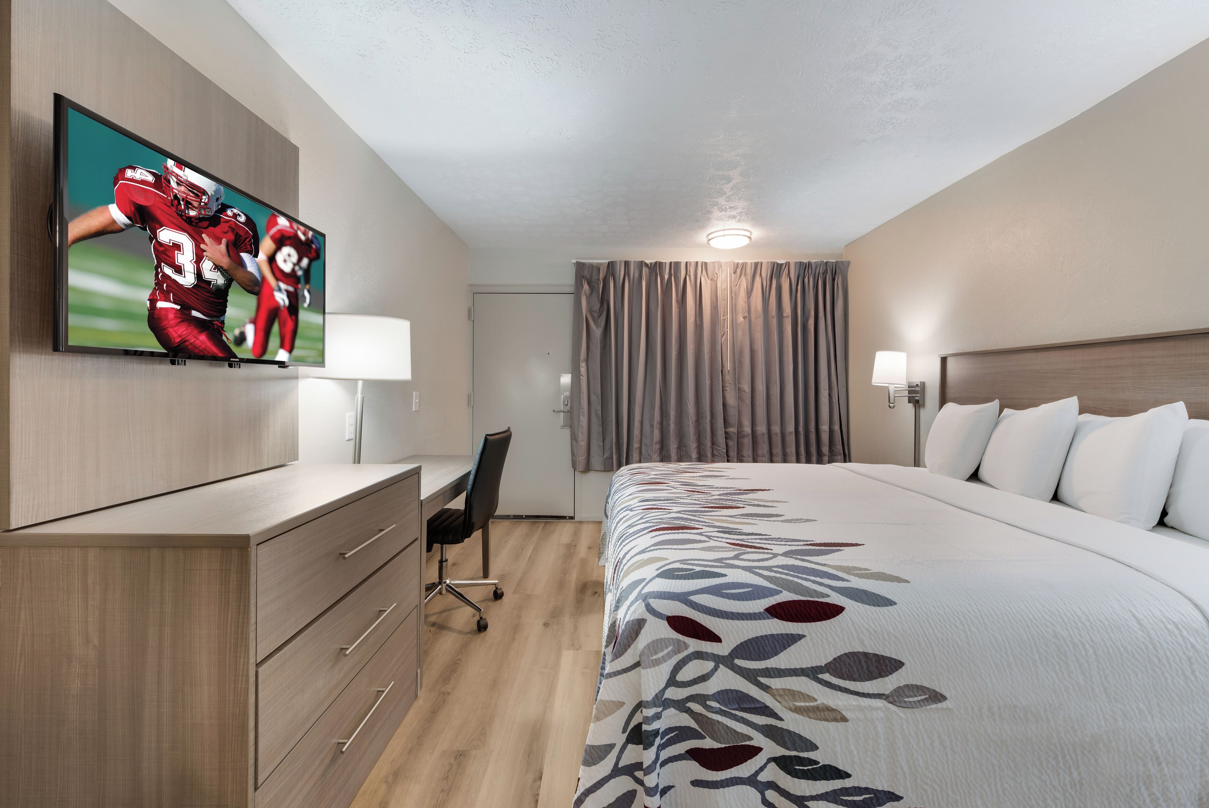 A hotel room with a large bed, a desk with drawers, a television mounted on the wall, a chair, a window with curtains, and a bedside lamp.