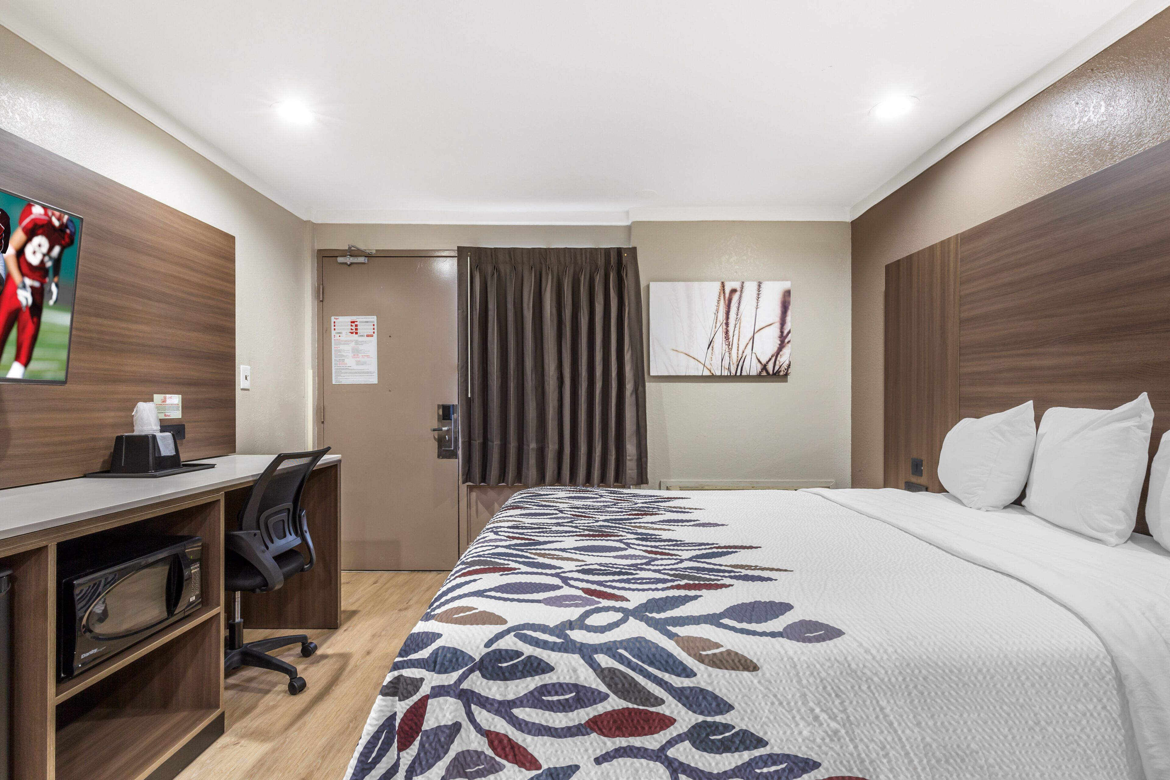 Une chambre d’hôtel avec un lit, un bureau, une chaise, une porte et un tableau accroché au mur.