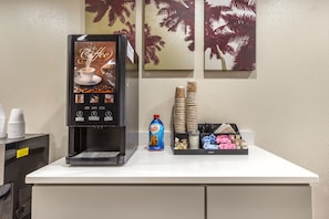 A coffee machine with a beverage selection panel, disposable cups, a bottle of coffee mate, and a tray with utensils and condiments. - Red Roof Inn Raleigh Northeast - Downtown (Raleigh)