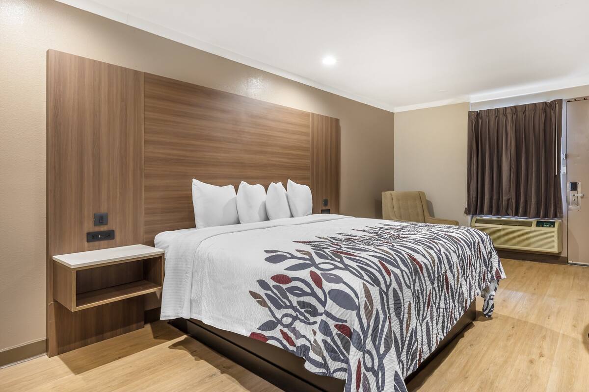 A hotel room with a wooden headboard, a bed with a floral-patterned bedspread, a bedside table, and a built-in shelf.