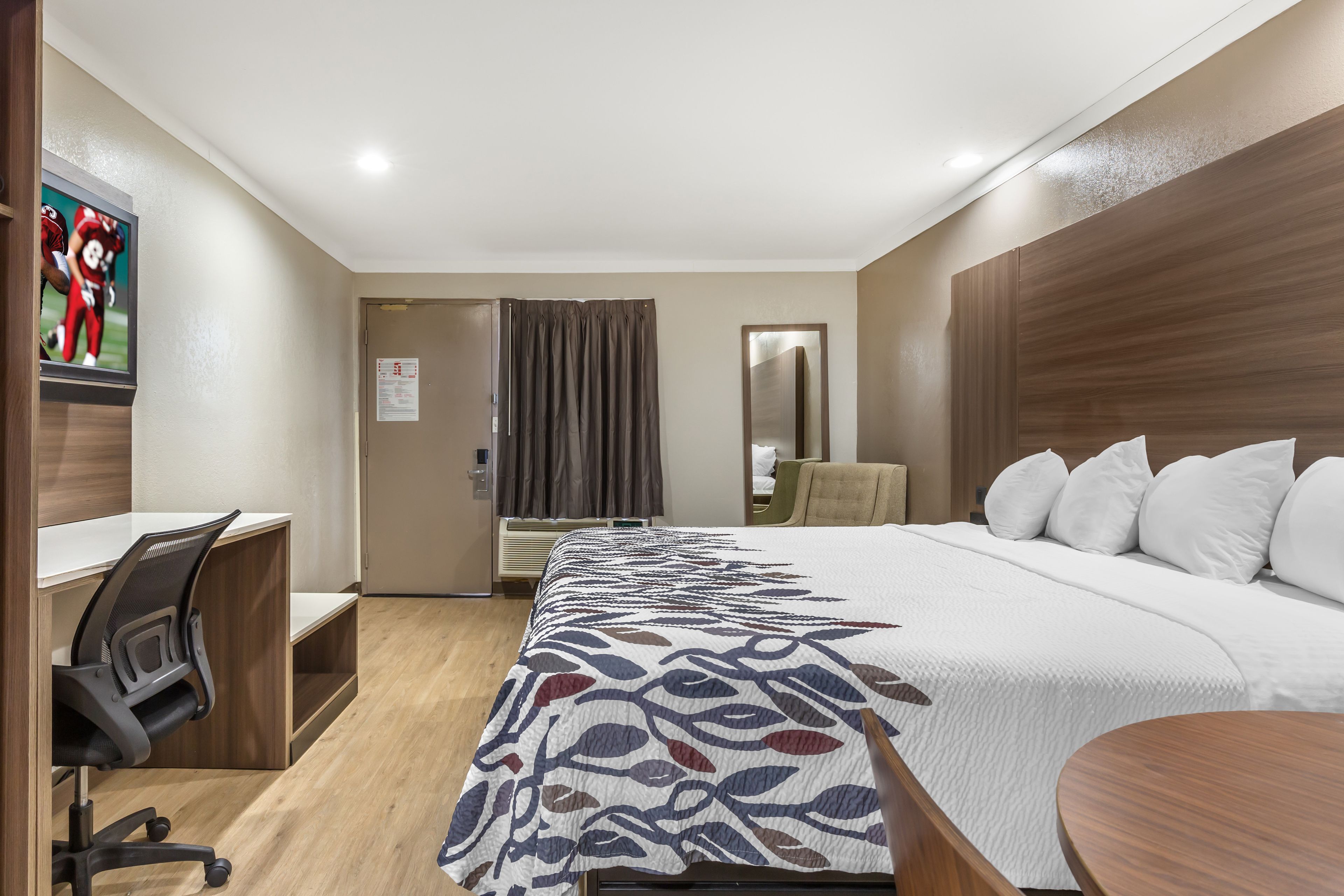 A hotel room with a bed, a desk, a chair, a mirror, and a television.