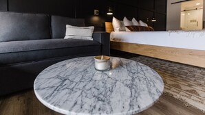 Una habitación de hotel moderna con un sofá gris oscuro, una mesa de centro de mármol redonda, una cama con cabecera de madera y una mesita de noche con lámpara.