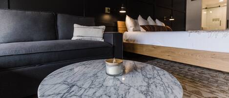 Une chambre d’hôtel moderne avec un canapé gris foncé, une table basse ronde en marbre, un lit avec une tête de lit en bois et une petite table de chevet avec une lampe.