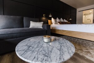 A modern hotel room with a dark grey sofa, a round marble coffee table, a wooden headboard bed, and a small bedside table with a lamp.