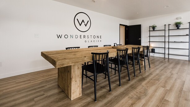 Un comedor moderno con una mesa de madera y sillas negras, una pared blanca con un logo, y una estantería negra con plantas.