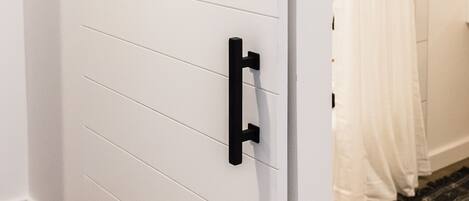 A white sliding door with black handles and a black track, leading to a bathroom with a white shower curtain.