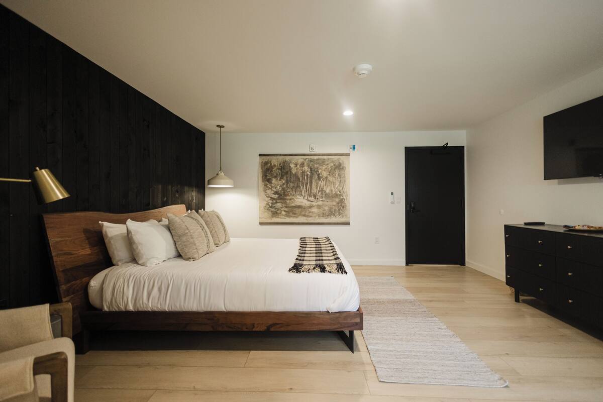 A bedroom with a wooden bed, a painting on the wall, a black wardrobe, and a television on the wall.
