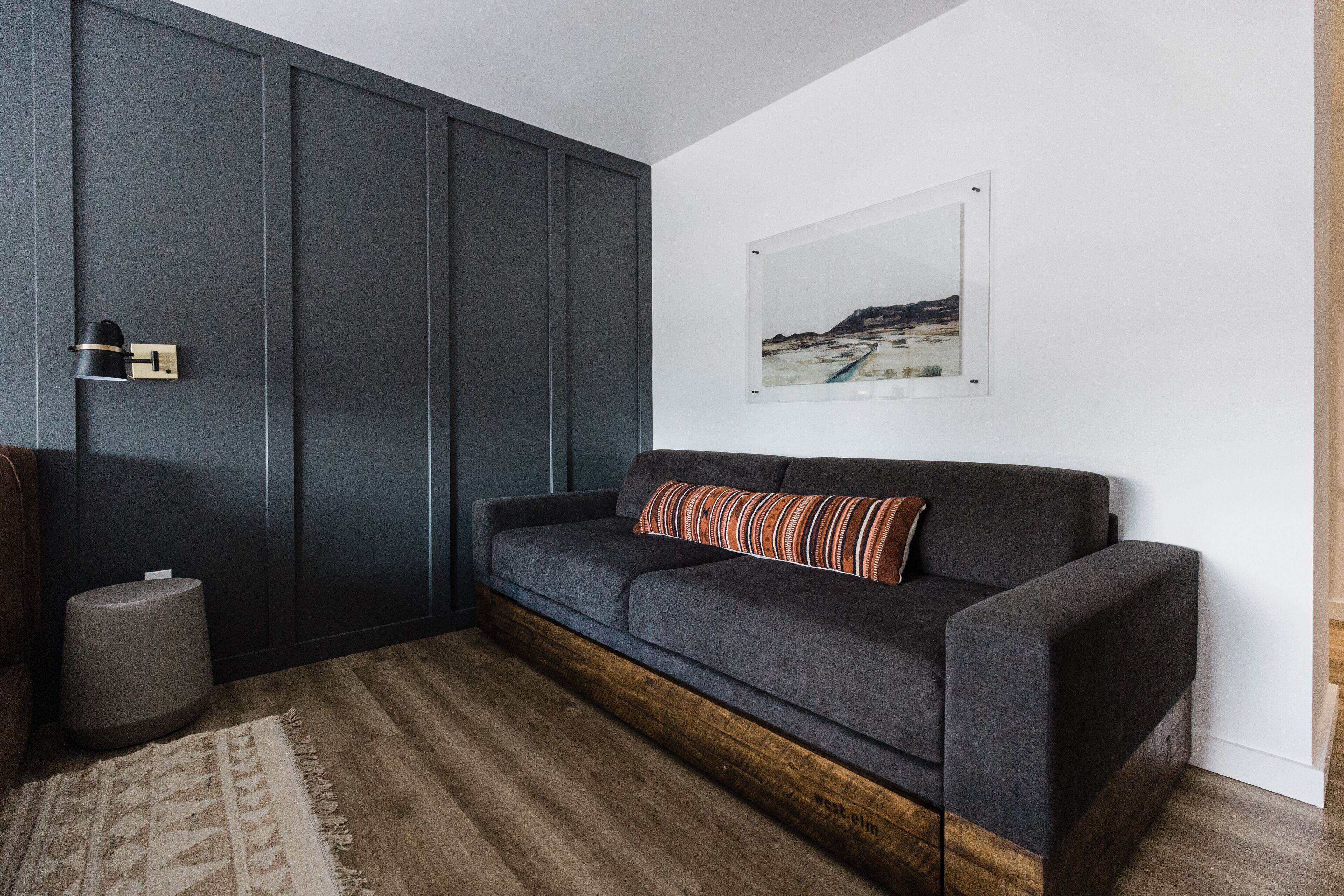 A modern living room with a dark grey sofa, a wooden floor, a small round ottoman, and a framed picture on the wall.