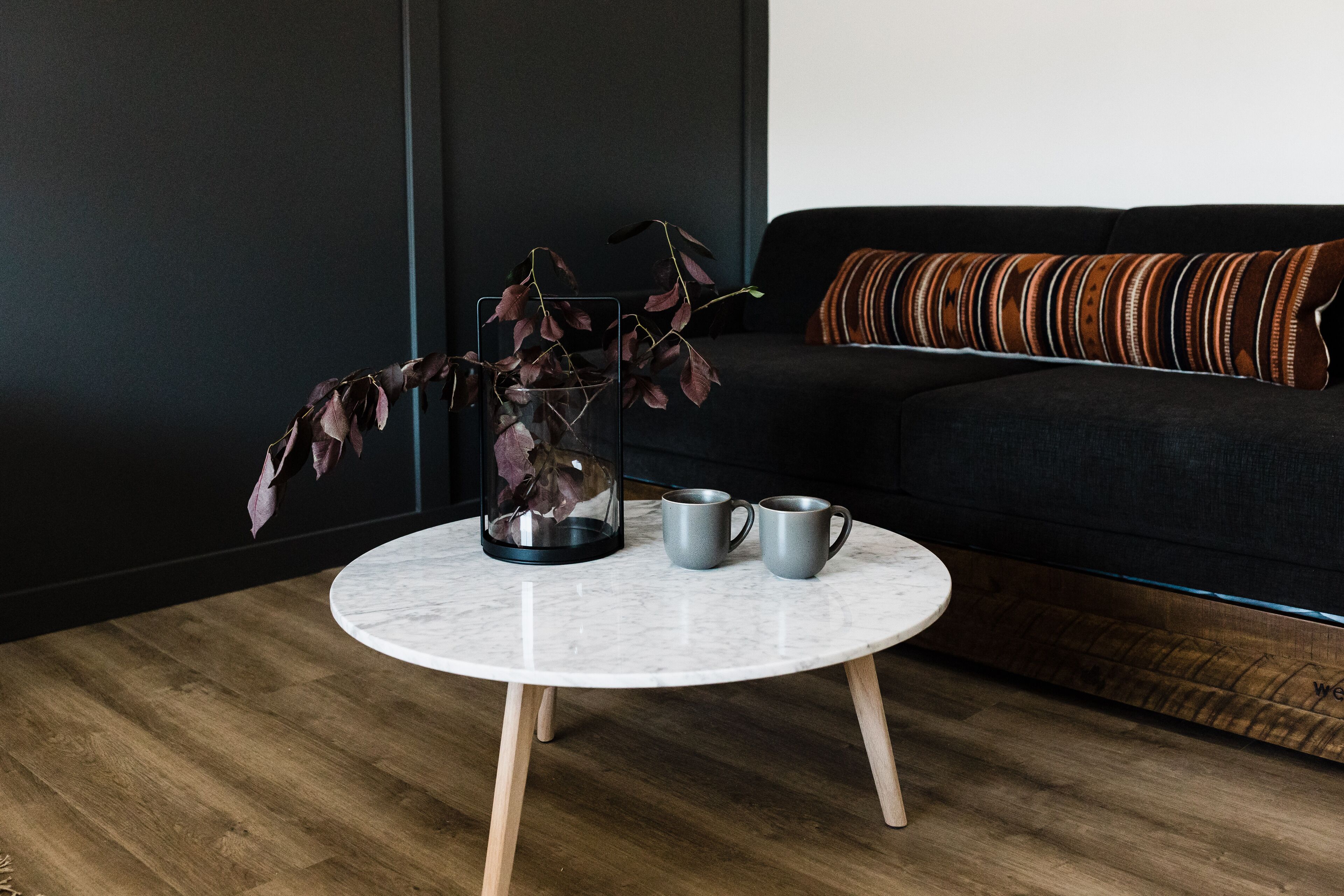 A round table with a glass vase and two mugs on a wooden floor, a dark sofa with a striped cushion, and a dark wall.