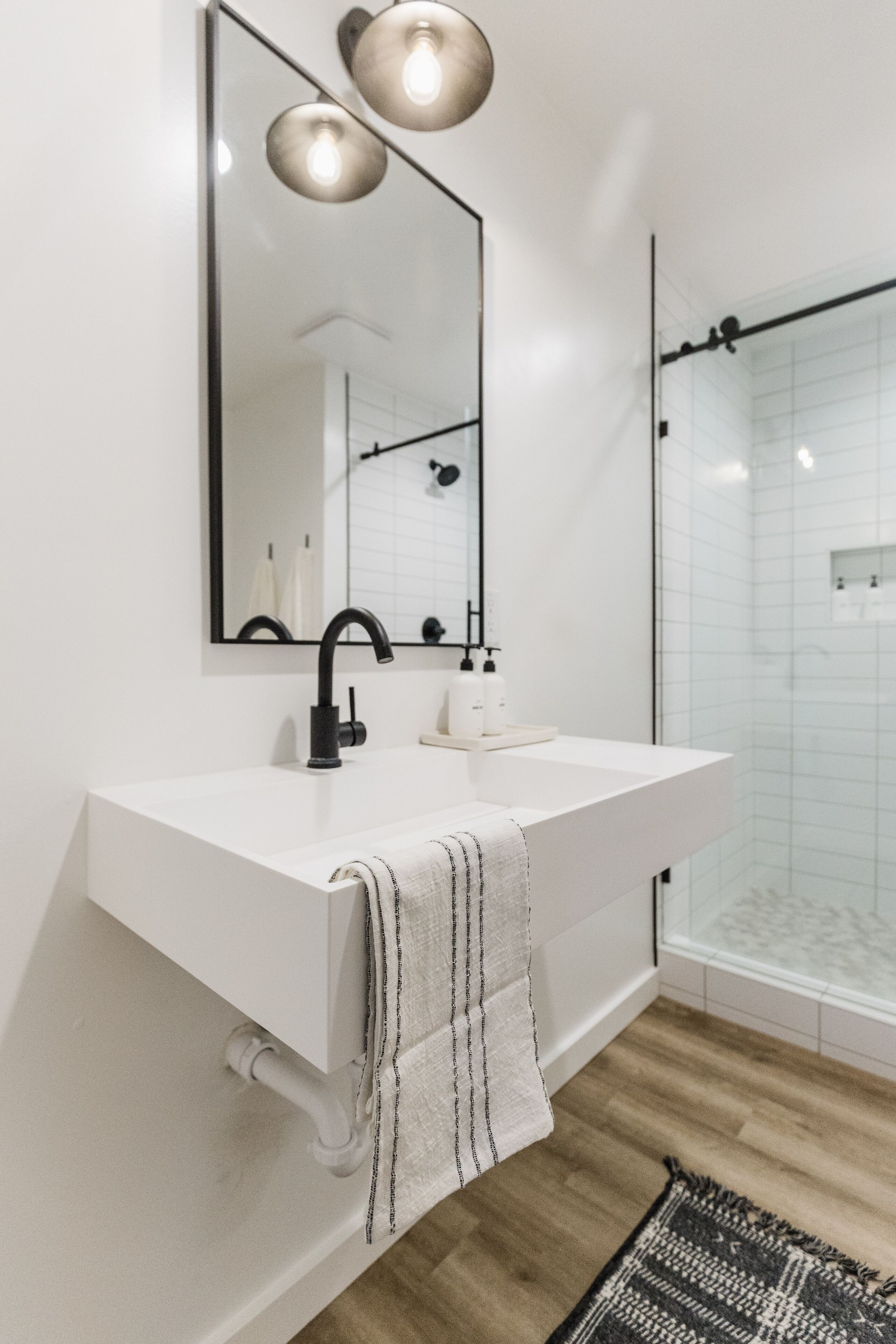 Un baño moderno con un lavamanos blanco, grifo negro y un espejo encima. Hay una toalla colgada en la pared y se ve una zona de ducha al fondo.