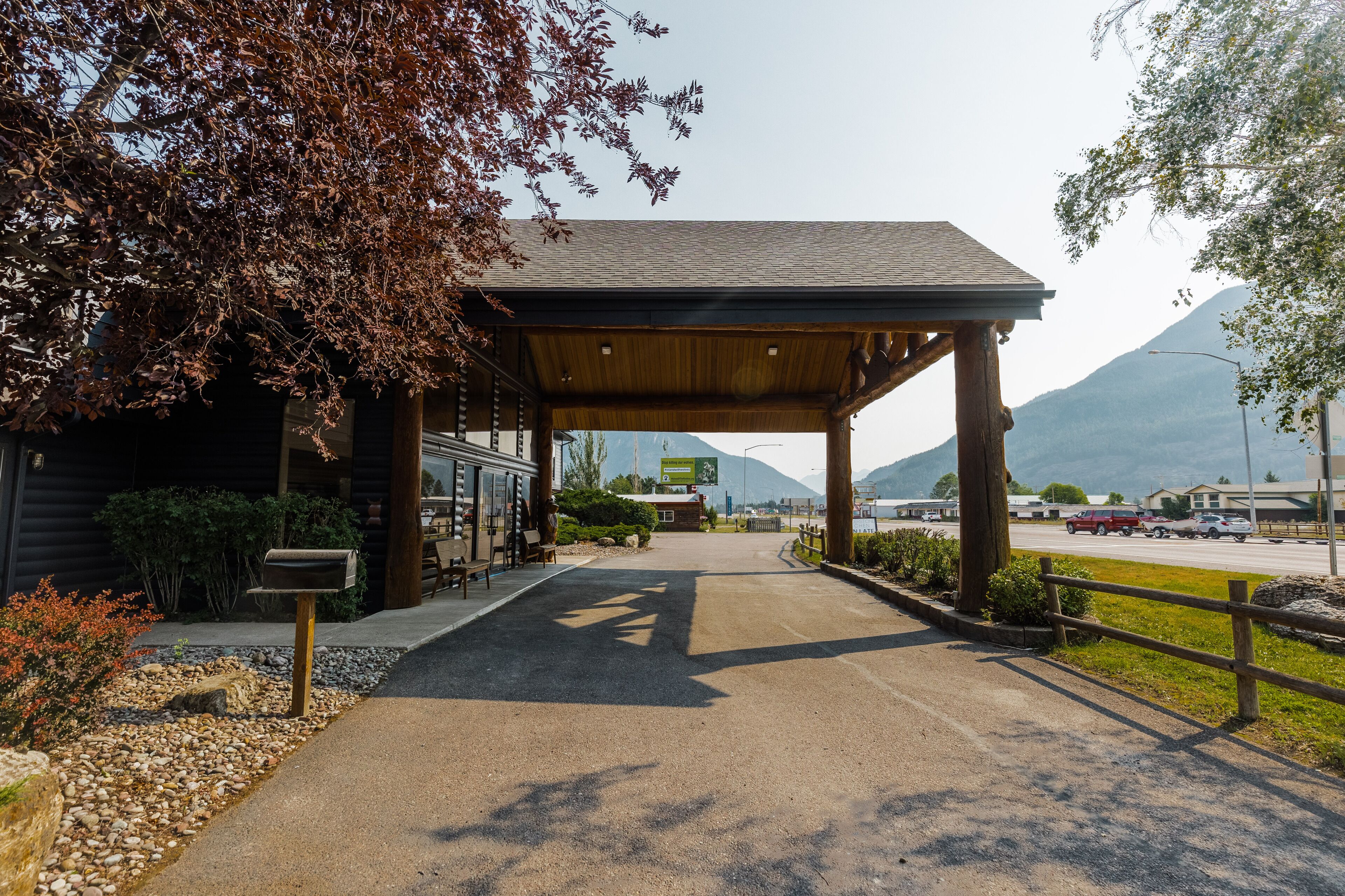 A covered entrance to a building with a gravel path leading to it, surrounded by trees and a mountain in the background.