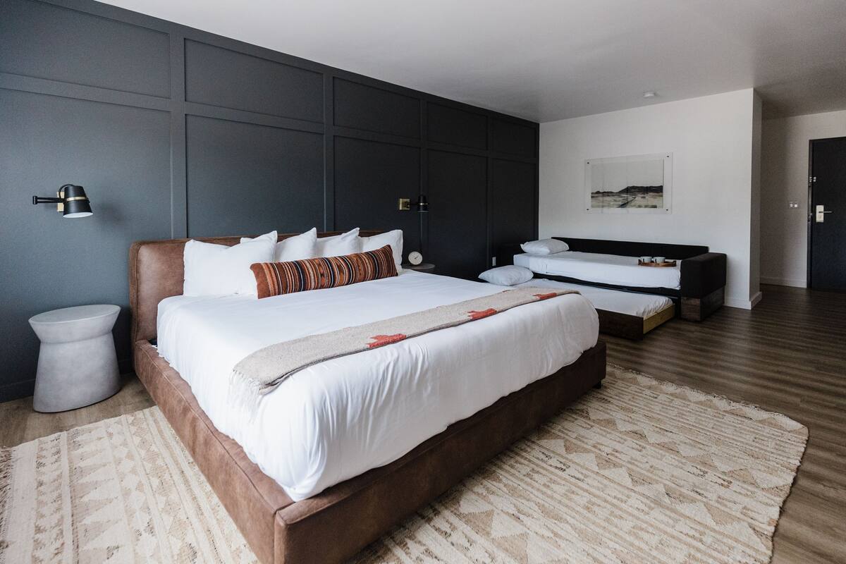 A modern bedroom with a large bed, a smaller bed, and a bench. There are two bedside tables and a wall-mounted lamp.