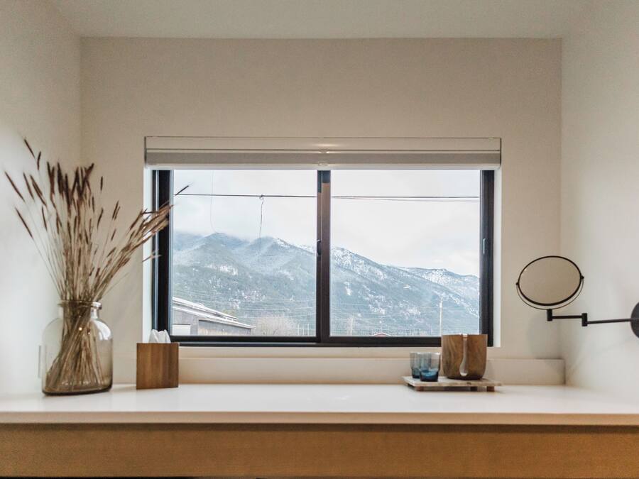 Une fenêtre donnant sur les montagnes, un vase contenant des plantes séchées et un petit miroir rond accroché au mur.