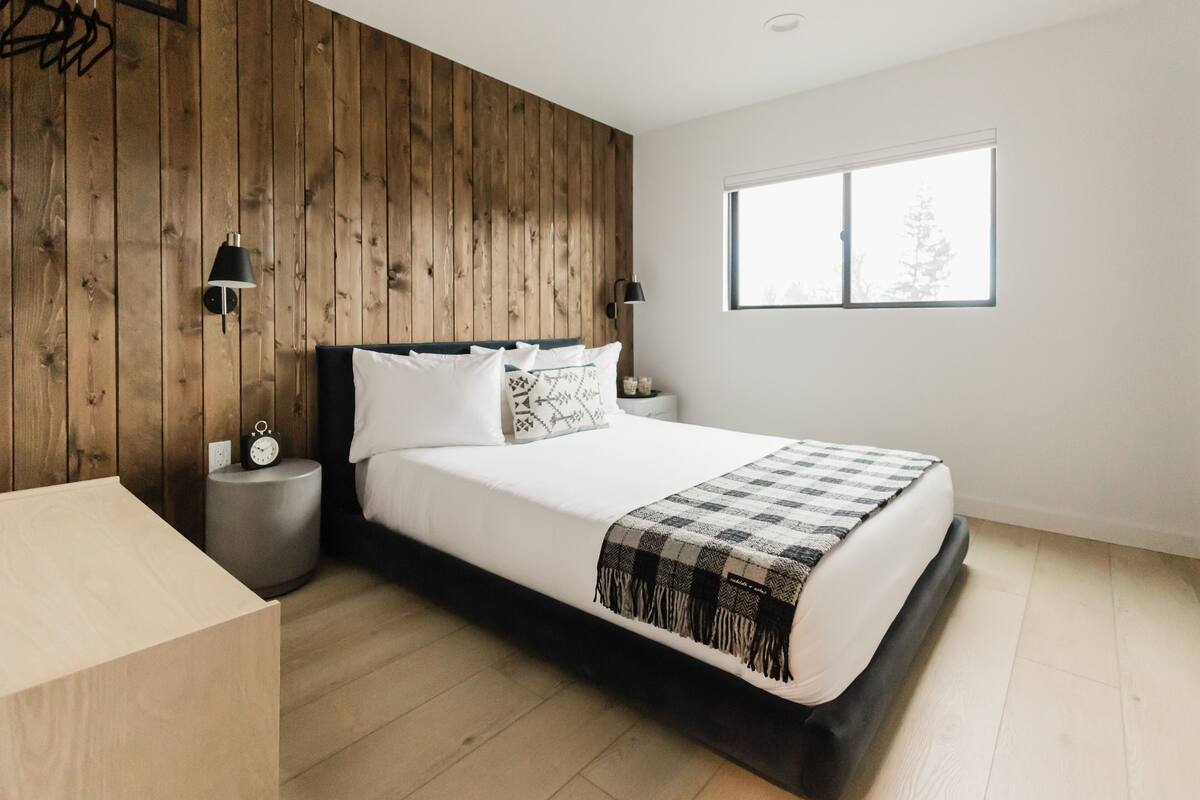 A bedroom with a wooden wall, a large bed with white bedding, two bedside lamps, a round nightstand with a clock, and a window with a view of trees.