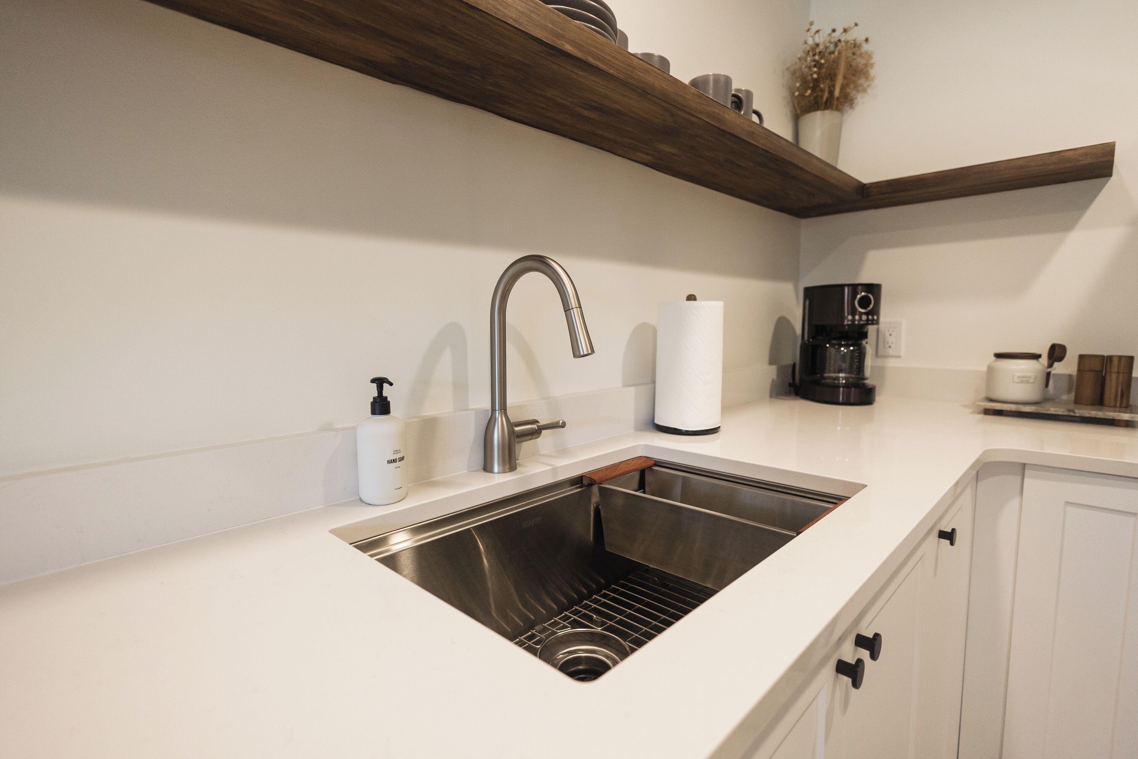 A modern kitchen with a stainless steel sink, white countertops, wooden shelves, and a coffee maker.